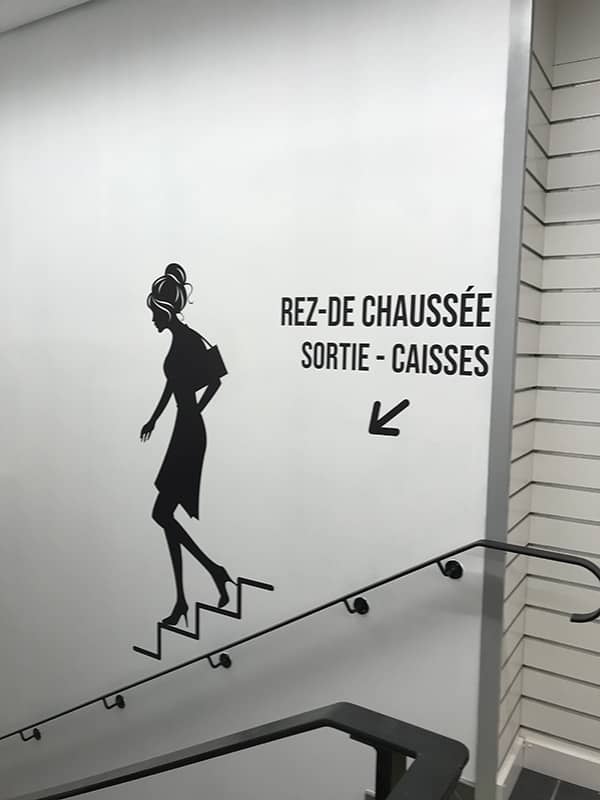 Signalétique directionnelle noir sur mur blanc avec dessin de femme descendant des escaliers
